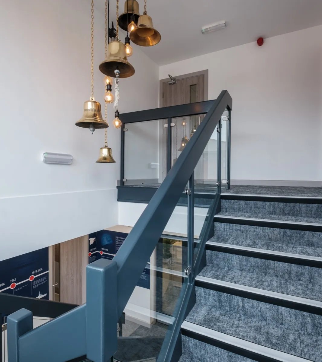 Staircase with cluster of brass ship's bell pendant lights and the Boluda history timeline display at Boluda Towage